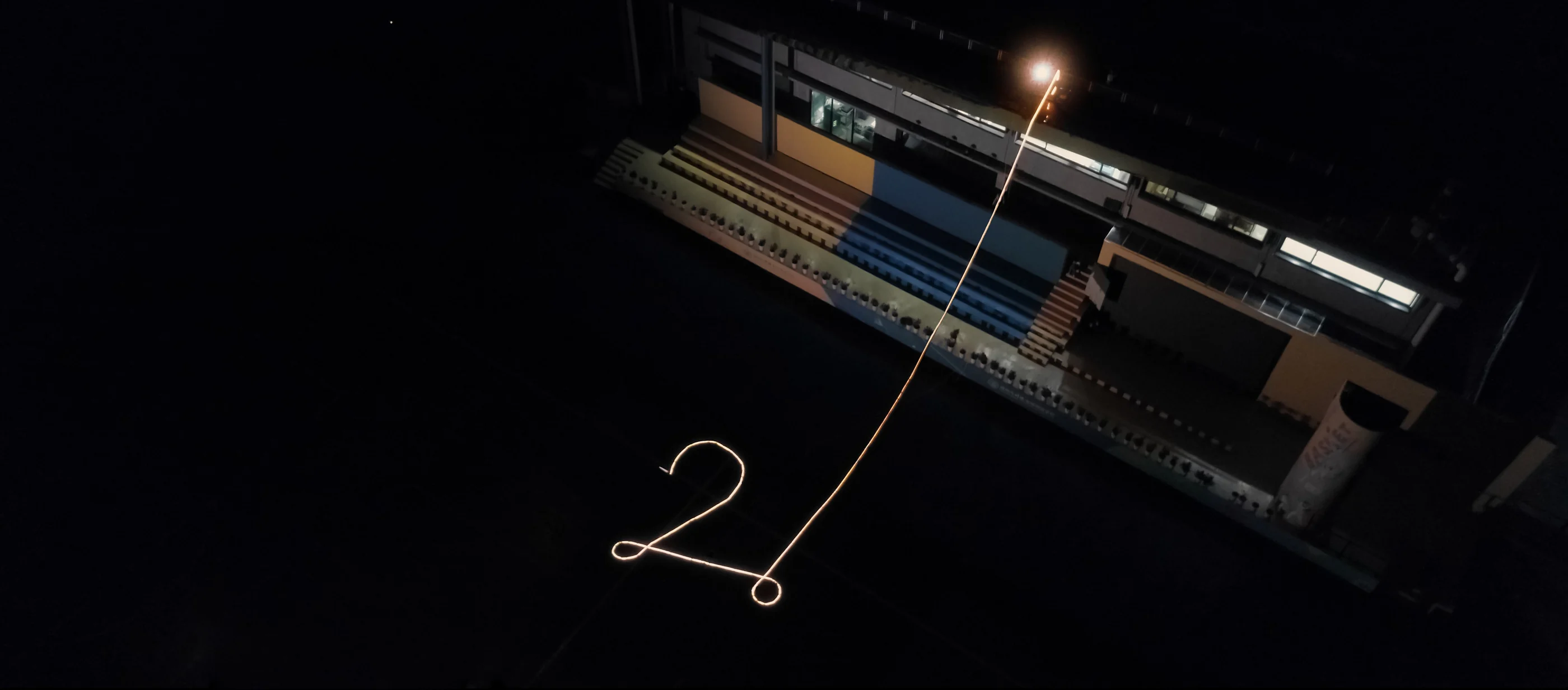 Aerial view at night: a glowing line drawn on the sports ground rises to a bright point on the roof of the school stand