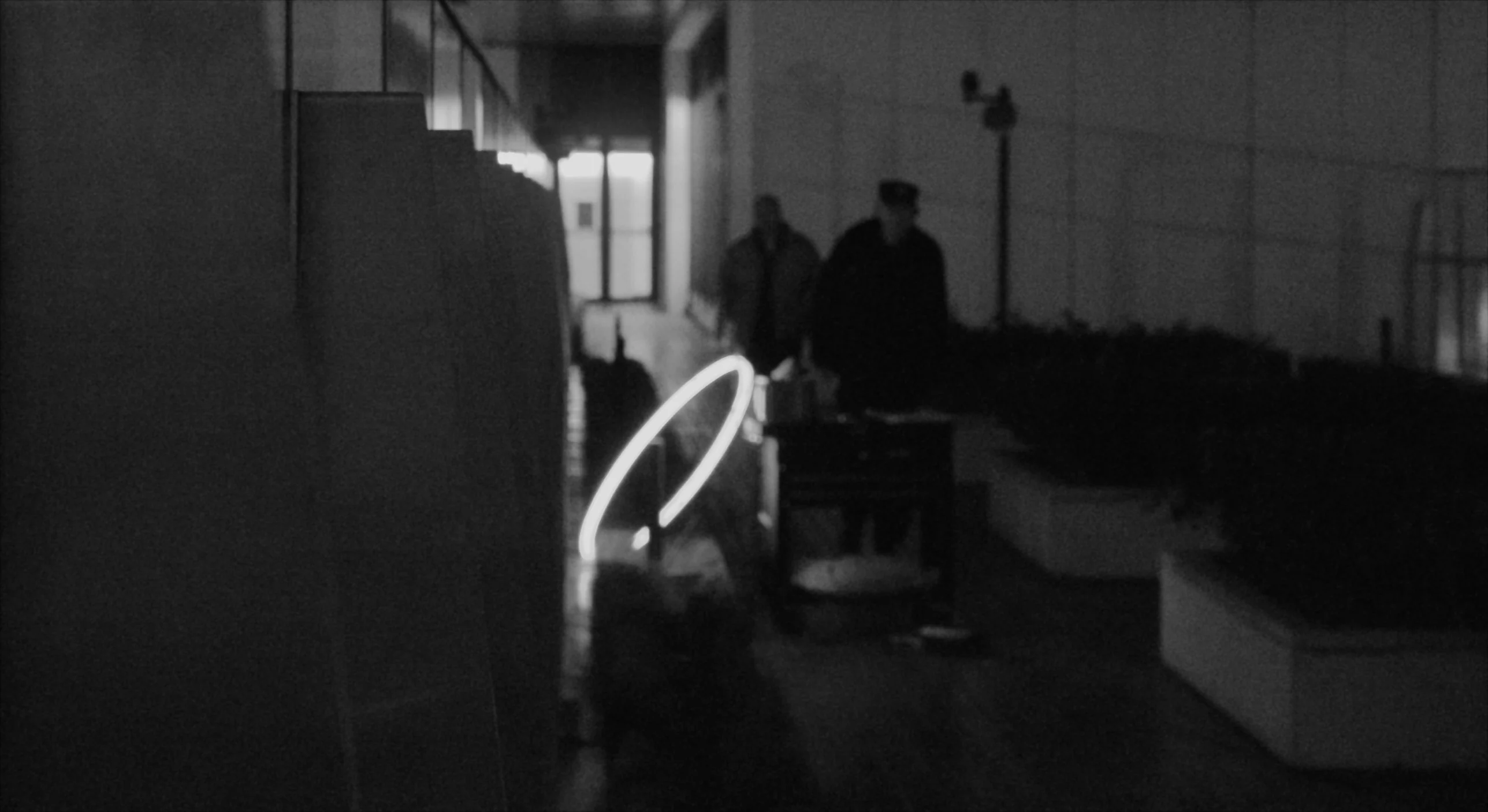 Black-and-white still: the glowing ring in a dim corridor as two security guards approach